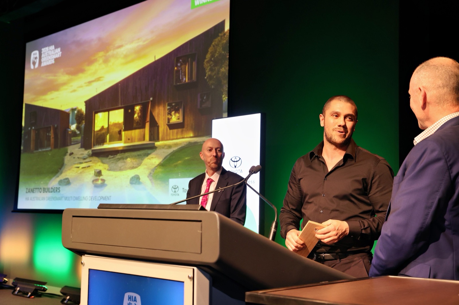 Zanetto Builders accepting a 2026 HIA Australian GreenSmart Award on stage in Melbourne, with the winning project shown behind
